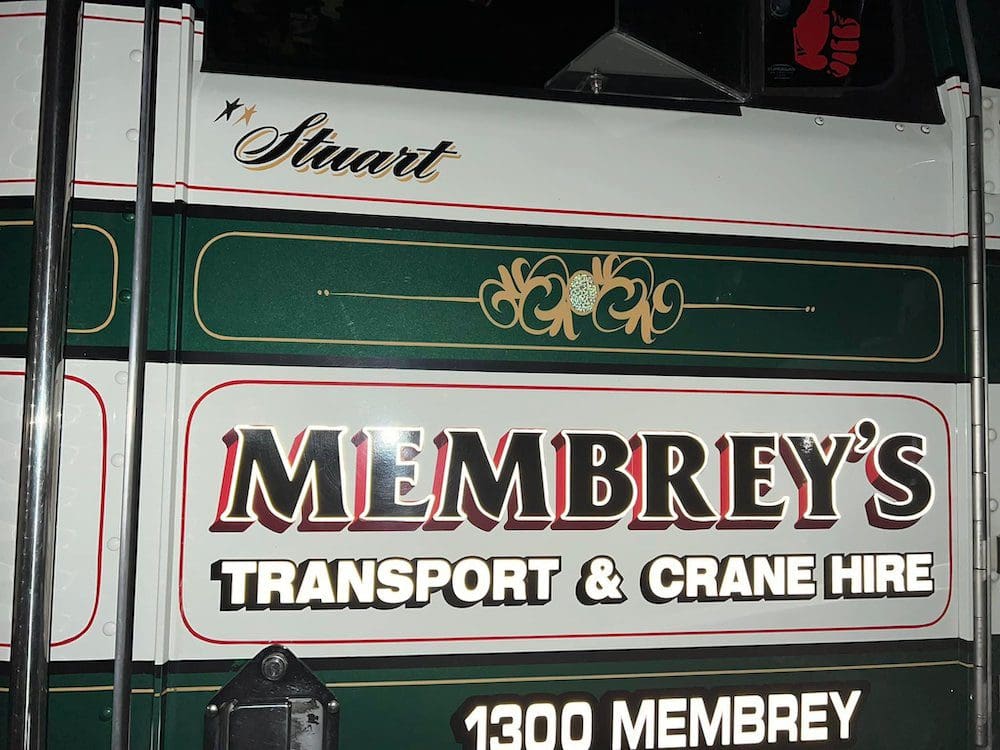 Side view of a truck door displaying the name "Stuart" above "Membrey's Transport & Crane Hire" in bold letters, with decorative gold detailing on a green and white background. The number "1300 MEMBREY" is also visible.