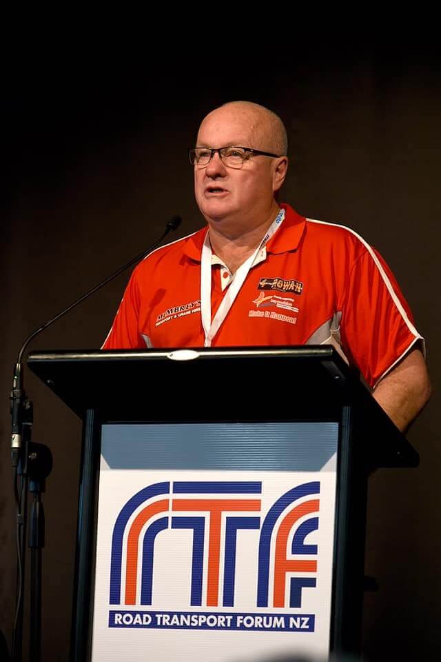 A man in glasses and a red polo shirt speaks at a podium with a microphone. The podium displays the logo and name "Road Transport Forum NZ" (RTF) on the front.