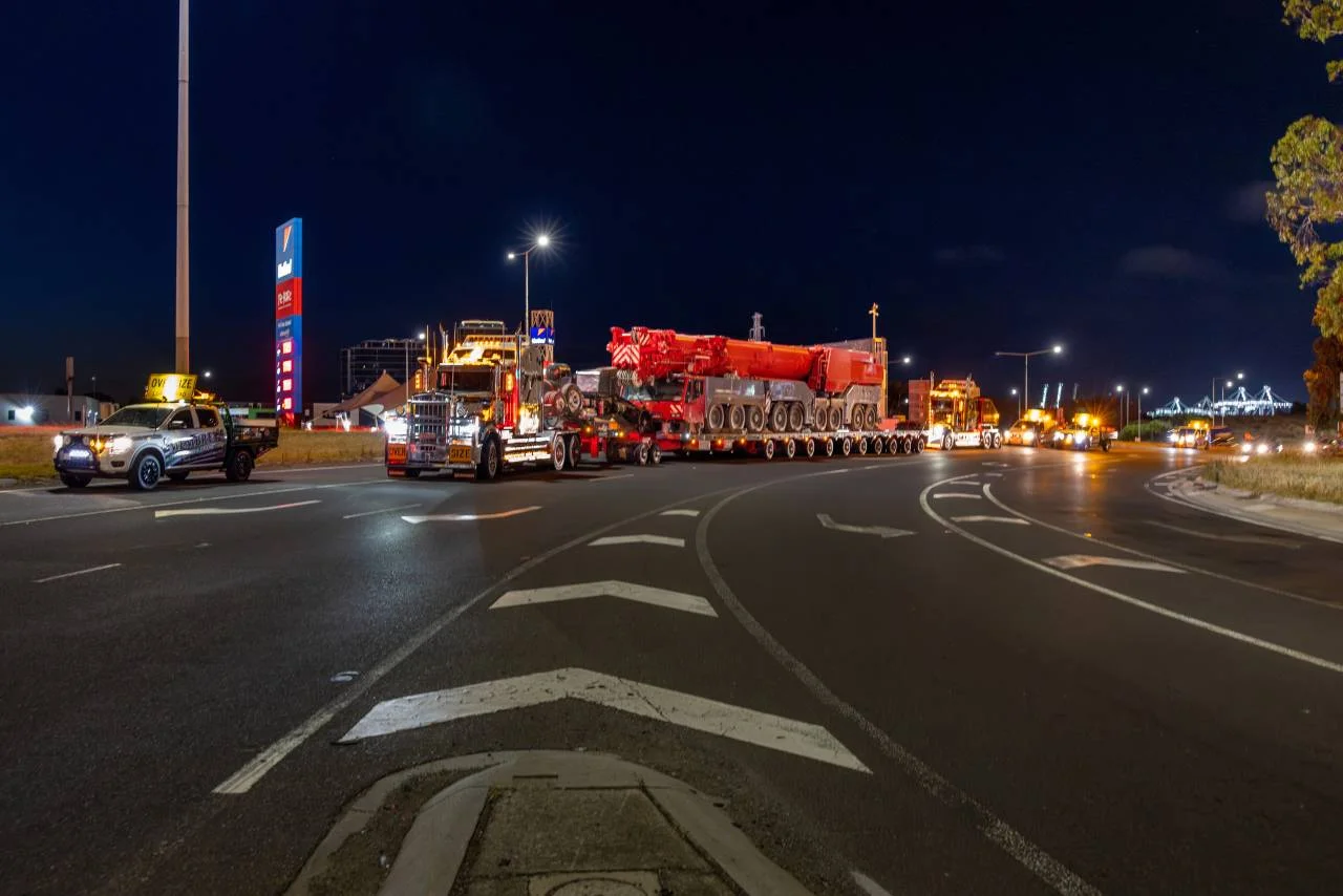A large truck with multiple axles transports an oversized red crane at night, escorted by several vehicles with flashing lights on a wide, well-lit road near a fuel station.
