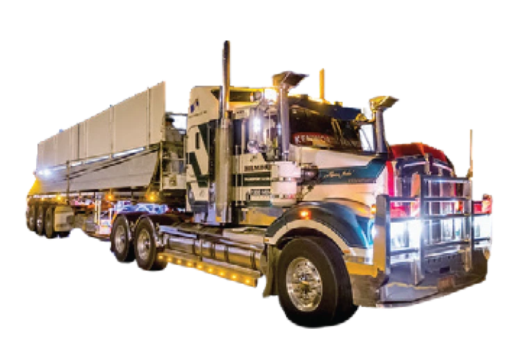 A large, multi-axle semi-truck with bright lights and a long flatbed trailer, transporting cargo at night. The truck has a shiny, polished exterior with chrome details.