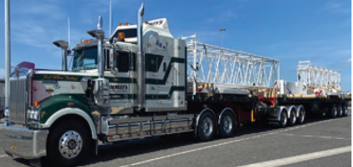 A large green and white semi-truck with multiple trailers is parked on a road, carrying long metal structures under a clear blue sky.