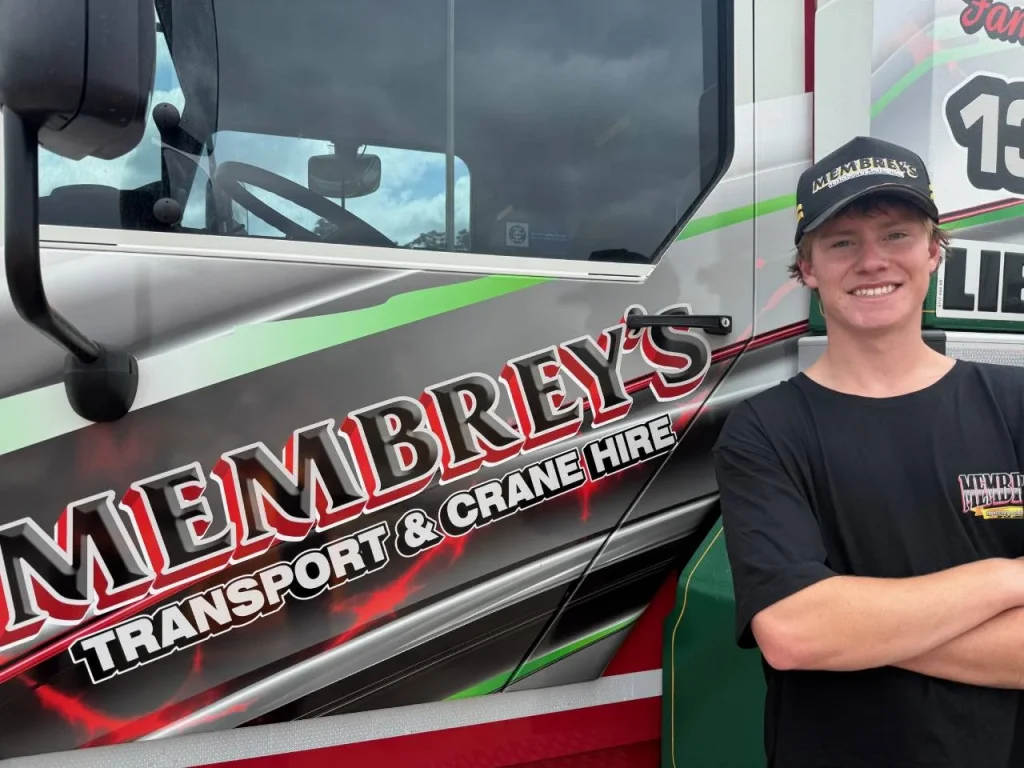 A young man in a black "Membrey's" shirt and cap stands smiling with arms crossed next to a truck displaying the "Membrey's Transport & Crane Hire" logo.