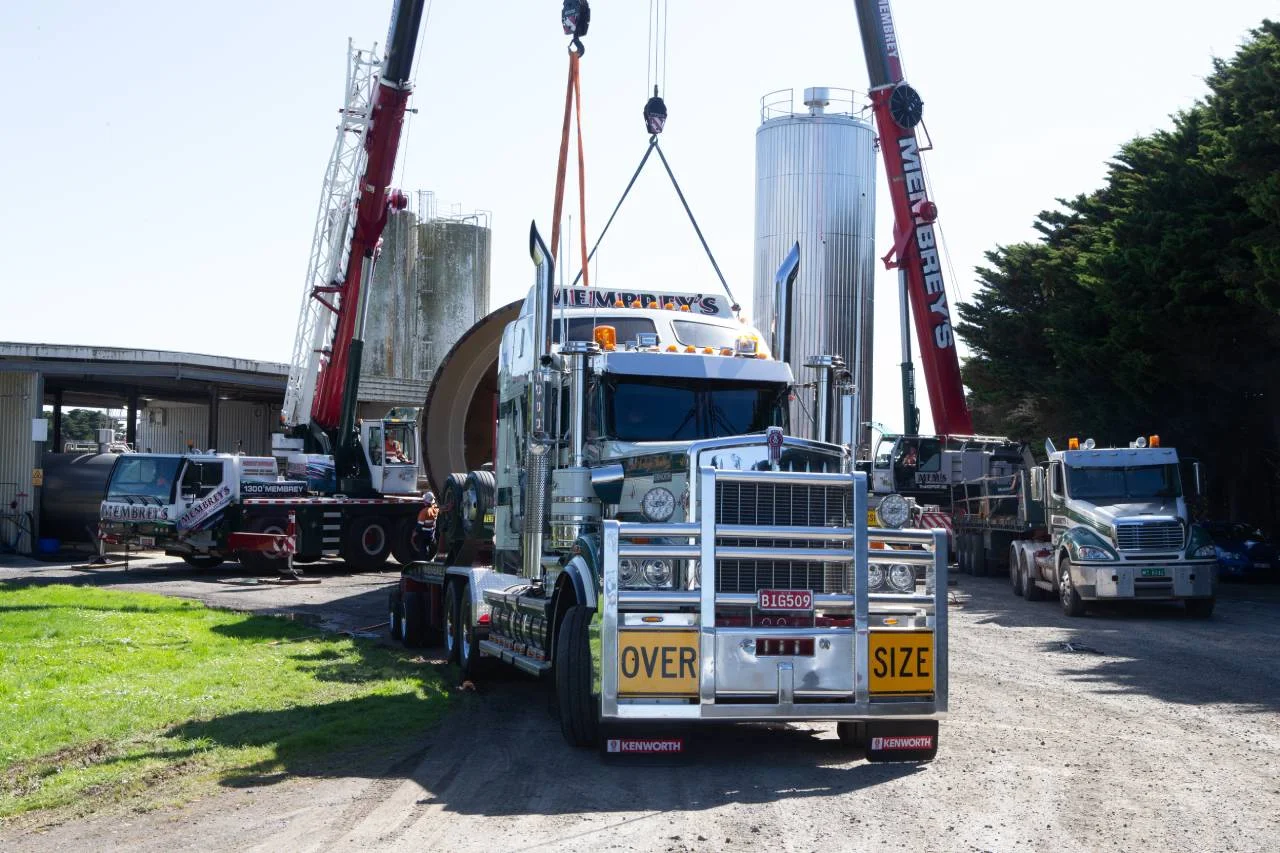 A large “Oversize” Kenworth truck is parked on a dirt lot as two cranes lift a massive cylindrical tank in the background. Other trucks and green trees are visible nearby under a clear sky.