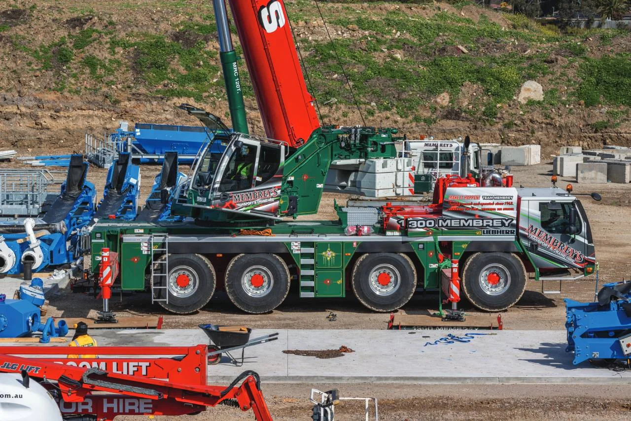 A large green and red mobile crane truck with “MEMBREY’S” branding is parked at a construction site, surrounded by various equipment and machinery on a concrete slab.