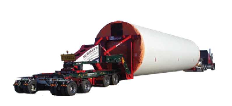 A large cylindrical wind turbine tower section is being transported on a specialized multi-axle trailer pulled by a truck. The tower section is white with an orange cover at one end.