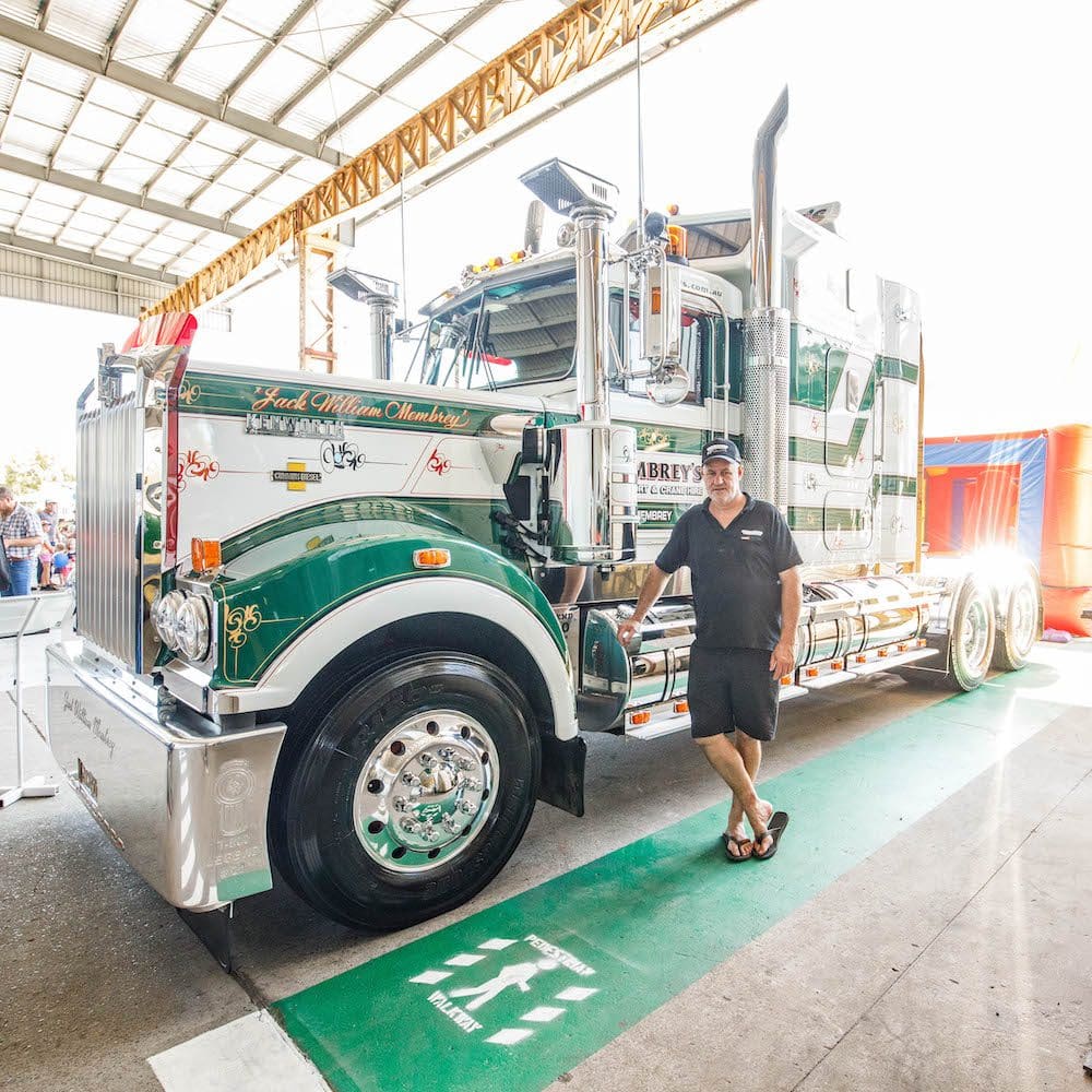 A man in a black shirt and cap stands smiling next to a large, shiny green and white semi-truck inside a well-lit building with a high metal roof.