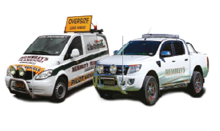 Two vehicles with "Kennedy’s" branding; one is a white van with an "OVERSIZE LOAD AHEAD" sign on the roof, and the other is a white pickup truck with green and gold detailing.