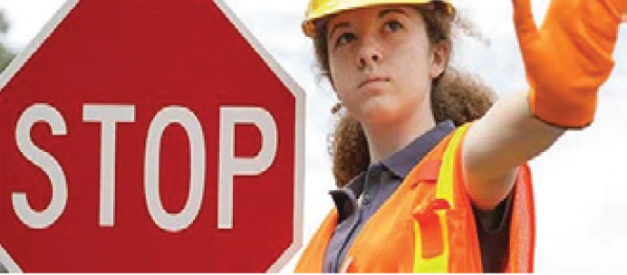 A person wearing a hard hat, orange safety vest, and gloves holds up their hand in a stopping gesture next to a large red stop sign.