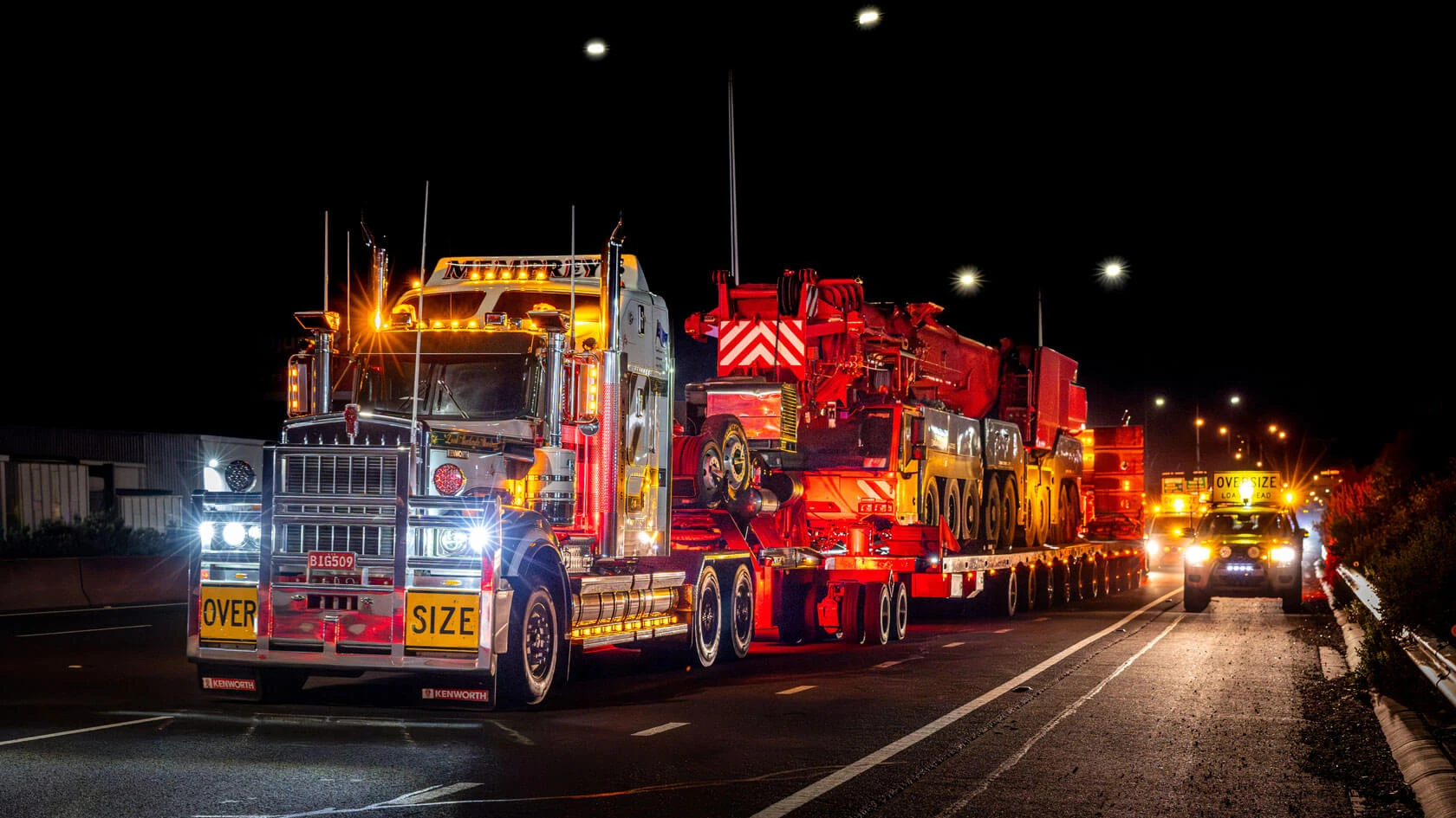 A large, illuminated truck labeled "OVER SIZE" transports heavy machinery at night, accompanied by escort vehicles with flashing lights on a roadway.
