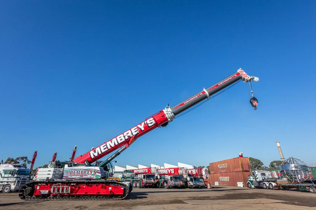 A large red and white crane with "Membrey's" written on its arm is extended upward at a construction site, surrounded by trucks, containers, and equipment under a clear blue sky.