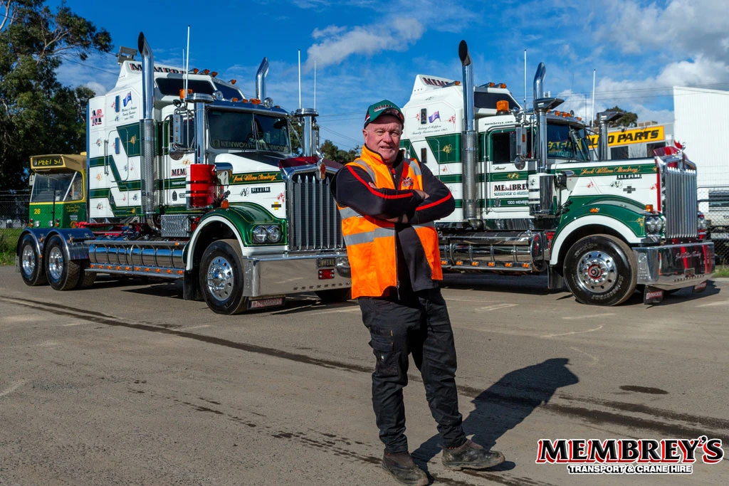 A man in an orange safety vest stands smiling with arms crossed in front of two green and white Membrey’s transport trucks. The Membrey’s logo is visible in the corner.