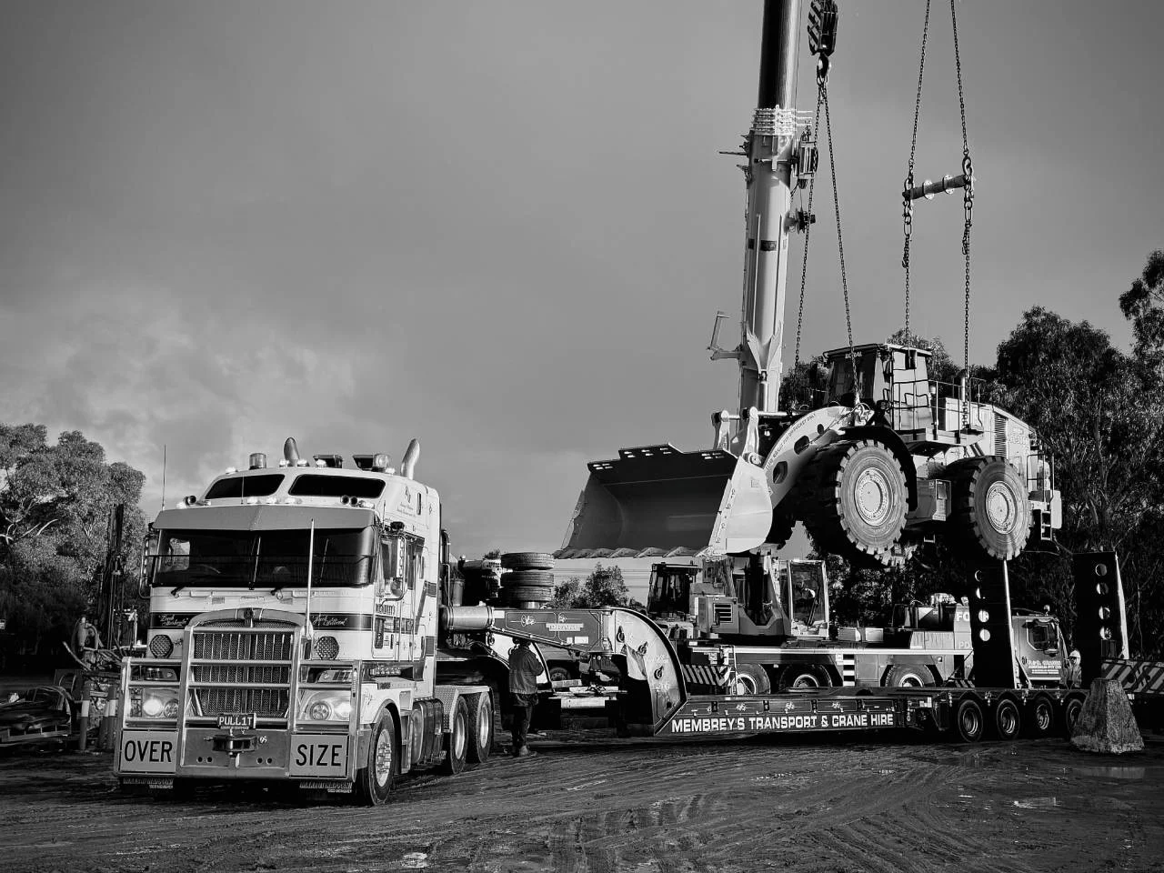 A large crane lifts a heavy mining vehicle onto a flatbed truck labeled "Oversize" and "Membrey’s Transport & Crane Hire" at a construction or mining site, with other equipment and trees in the background.