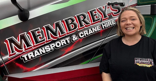 A smiling woman wearing a Membrey’s Transport & Crane Hire T-shirt stands in front of a truck displaying the company’s logo in bold red and white letters.