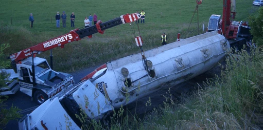 A large tow truck uses a crane to lift a toppled tanker truck from a roadside ditch while several people observe from a grassy field nearby.