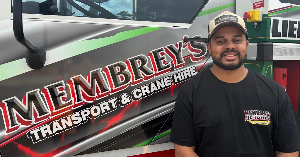 A man wearing a black Membrey’s t-shirt and a cap stands next to a truck with the Membrey’s Transport & Crane Hire logo on the side.