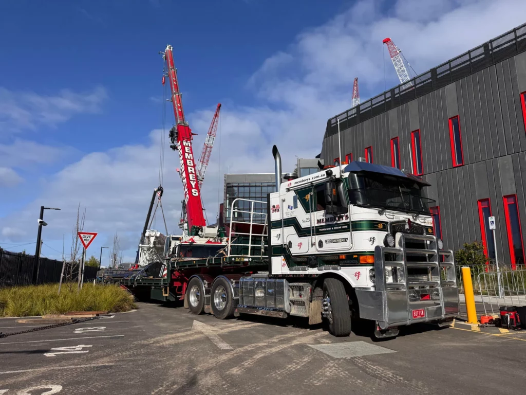A large white and green semi-truck is parked near a construction site with a red crane in the background. Modern buildings with red window frames and several cranes are visible under a partly cloudy sky.