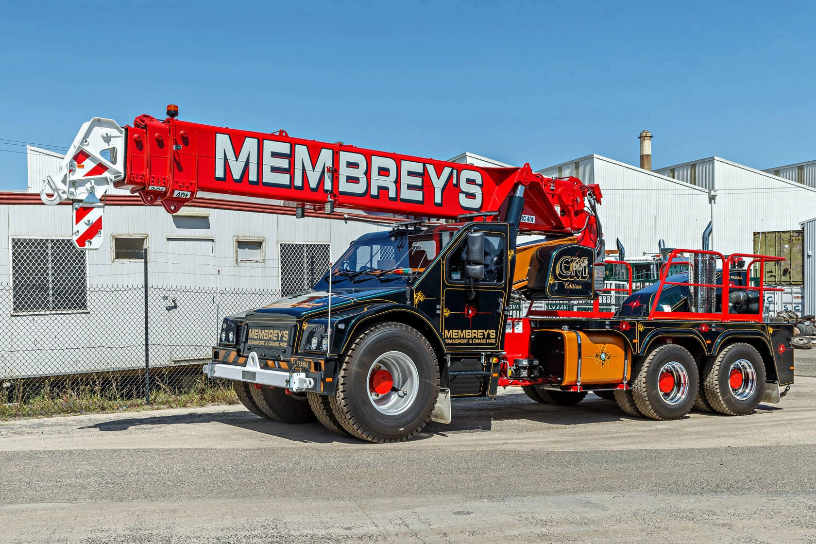 A large mobile crane truck with "MEMBREY'S" written on the extended red crane arm, parked outside industrial buildings on a sunny day.
