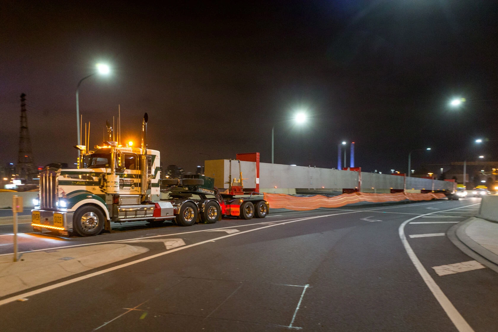 A large truck with multiple axles transports an oversized, long load at night, accompanied by bright streetlights on a city road with industrial structures faintly visible in the background.