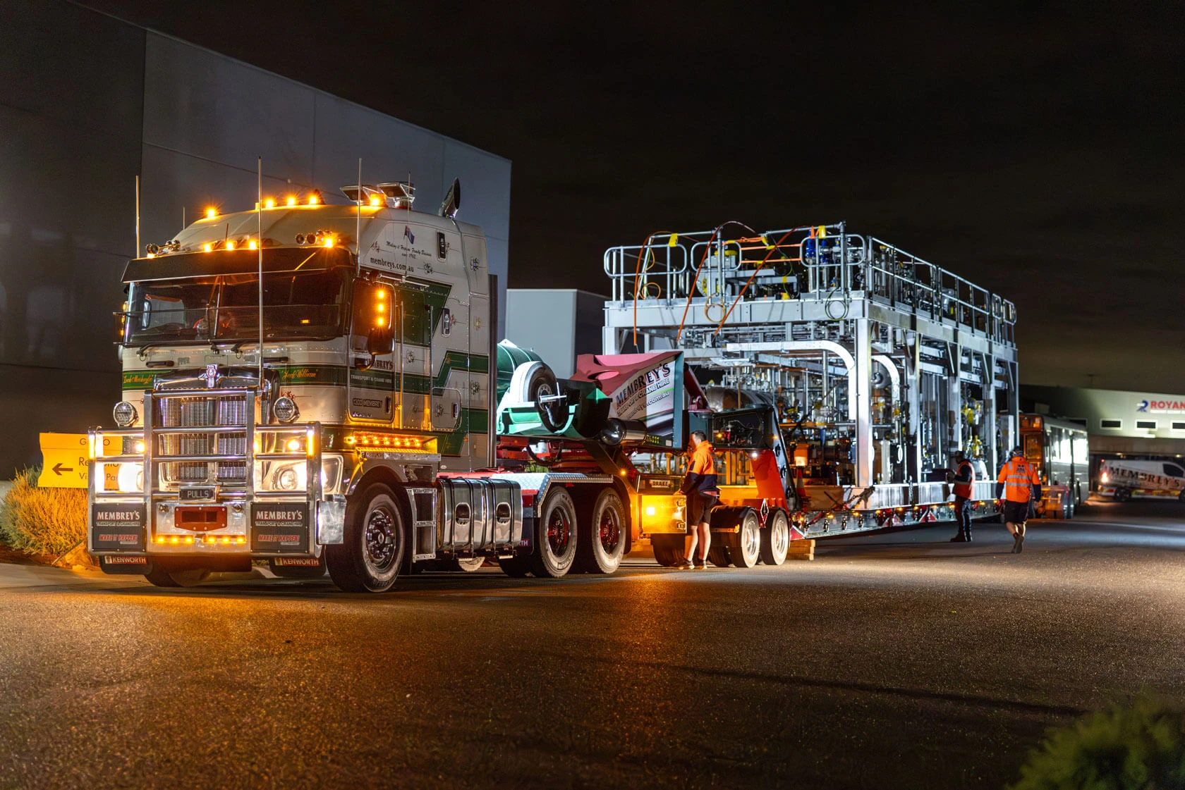 A large illuminated semi-truck transports heavy industrial equipment at night. Workers in high-visibility gear guide the loading process outside a commercial building.