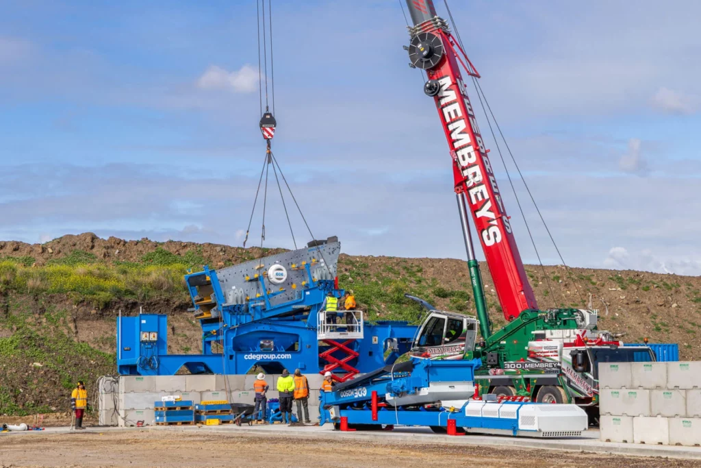 A large red crane marked "NEMBREYS" lifts industrial machinery at a construction site, with workers in safety gear nearby and a blue machine labeled "cdagroup.com" on the ground.