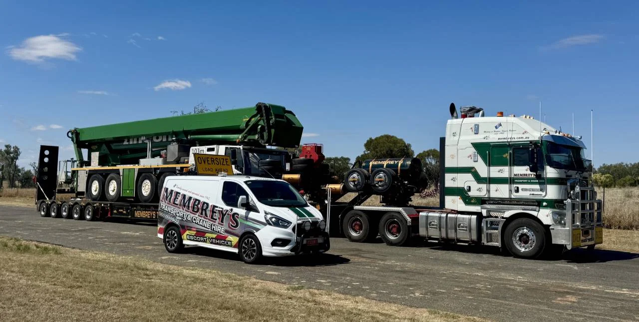 A large white and green semi-truck hauls heavy machinery on a flatbed trailer. In front, a white van with "Membrey's" and "Oversize Load" signs is parked on a rural road under a clear sky.