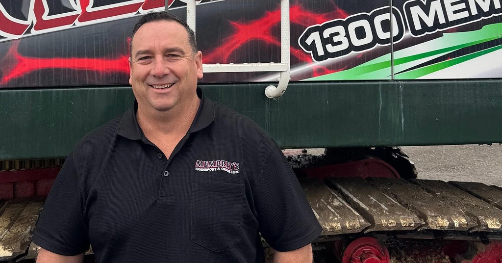A smiling man in a black polo shirt stands in front of a large vehicle with visible treads and colorful signage featuring the number "1300" and the word "MEM".