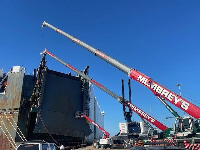 Three large red cranes labeled "Embrey’s" lift or support the partially opened ramp of a massive cargo ship at a dock, with a clear blue sky in the background. Other vehicles and equipment are nearby.