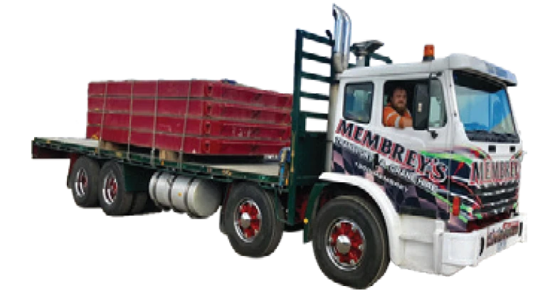 A flatbed truck with "Membrey's" branding carries stacked red pallets. A person wearing an orange vest is seated in the driver's seat, looking out the window.